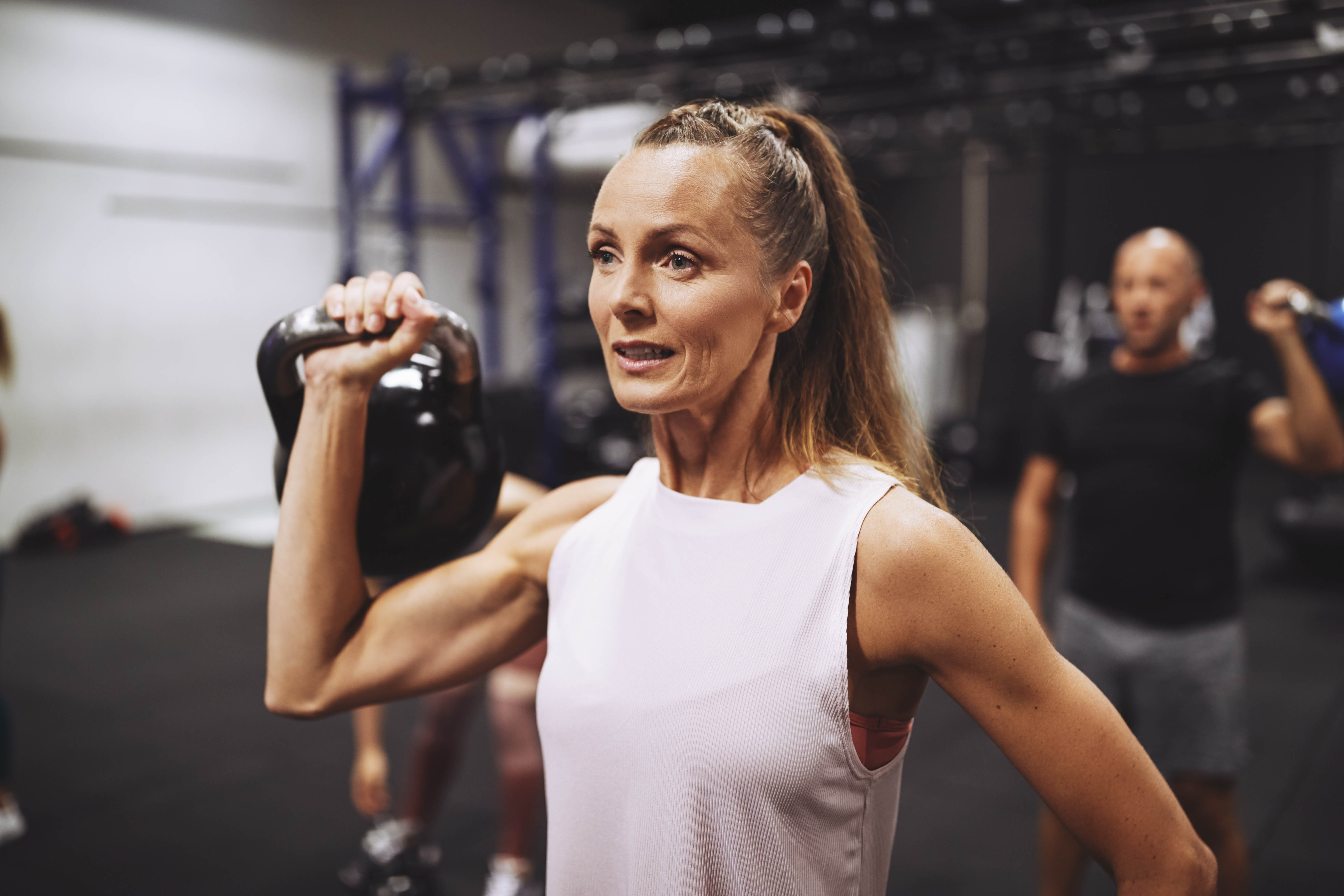 Strong, middle aged woman standing in a gym mid-workout, with a kettlebell in her hand at her shoulder height